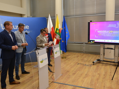 Uczestnicy konferencji przy mównicach na tle ścianki promocyjnej z napisem Lubuskie. Z boku telewizor.