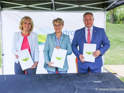 Trzy osoby stoją za stołem i trzymają w rękach teczki.