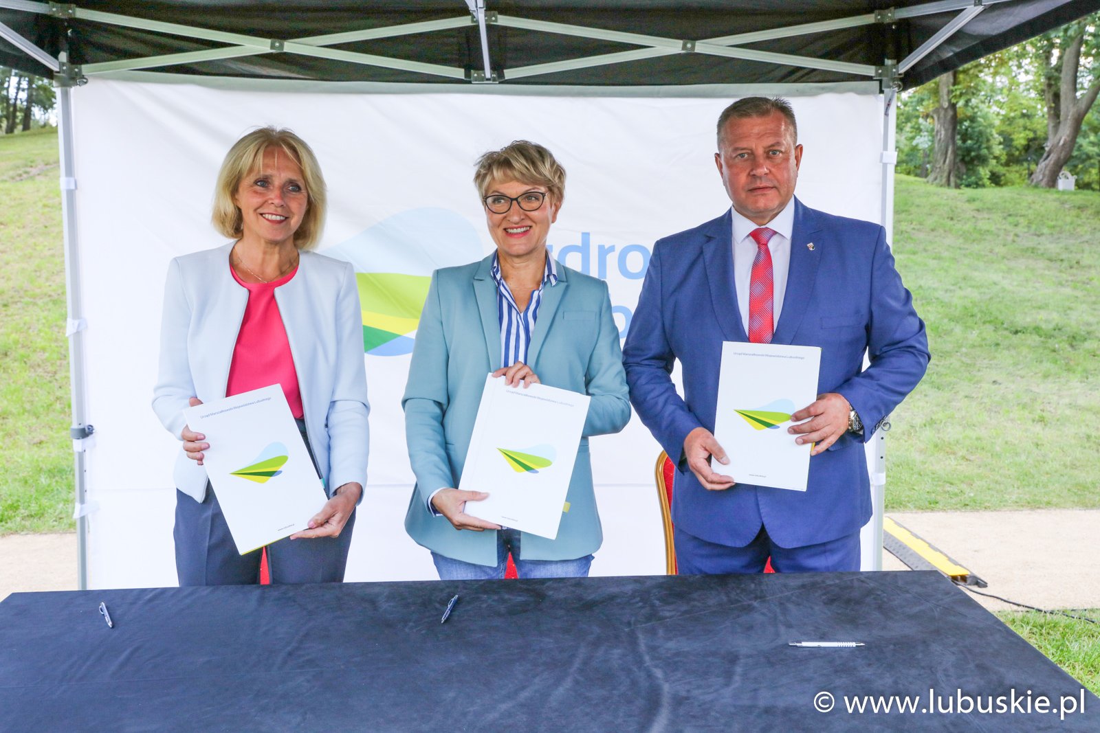 Trzy osoby stoją za stołem i trzymają w rękach teczki.